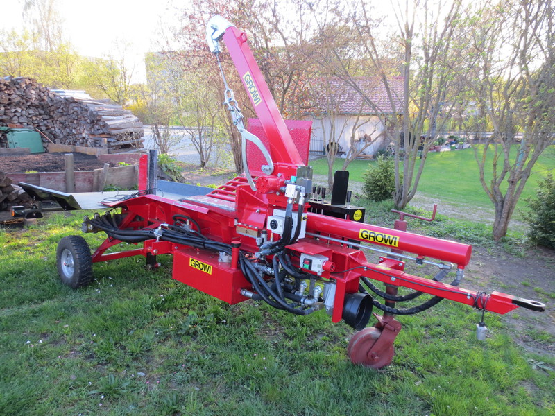 Roter Holzspalter-Anhänger im Garten vor einem Holzstapel, im Hintergrund sieht man ein Einfamilienhaus